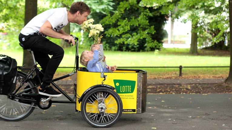 Mann fährt ein Lastenrad, in dem zwei Kinder sitzen